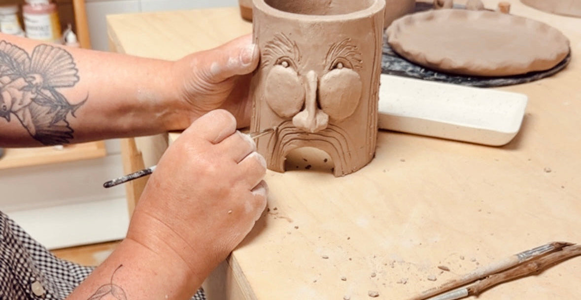 A close-up of a person's tattooed hands carving a clay cup with a stylized face on a wooden pottery workbench.