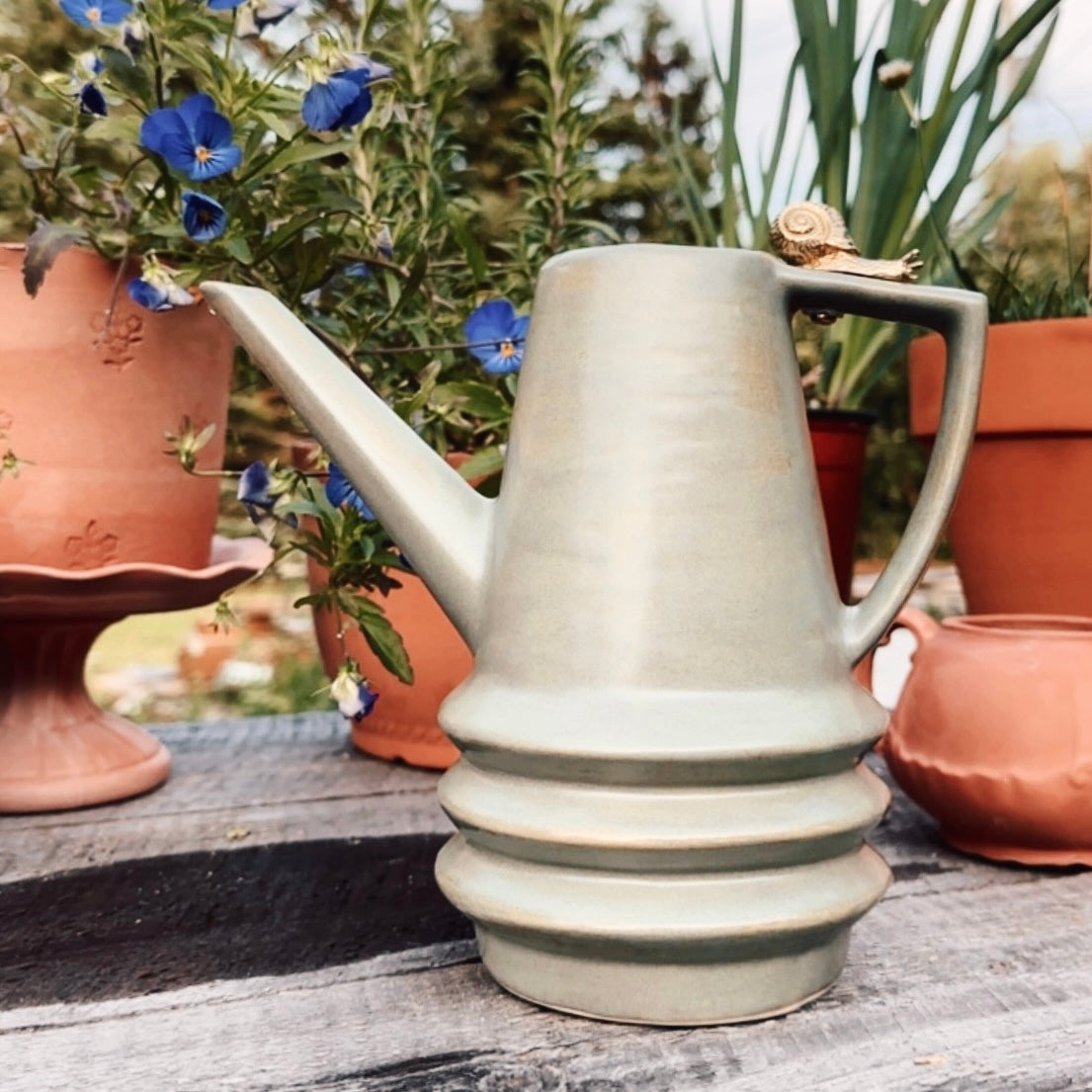 A light sage green ceramic pitcher with a textured finish, featuring a small gold snail detail on the handle, set on a rustic wooden surface with potted plants in the background.