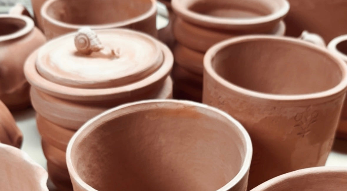 A collection of handmade terracotta clay pots and jars with matching lids, arranged closely together on a light surface. The pots feature natural hand-painted floral embossing and a warm, earthy reddish-brown color.